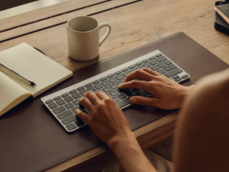 Mani al lavoro su una scrivania ordinata, con tastiera, quaderno e tazza